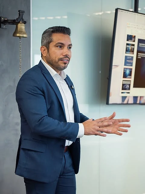Thiago Farias, de terno, apresentando em pé num escritório